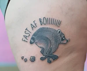 Черно-серая татуировка на коже, изображающая бегущего енота с текстом FAST AF BOIIIIII!! над ним