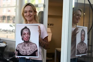 Женщина держит портрет с изображением своего лица и тела, покрытых принудительными татуировками, символизируя травму и процесс восстановления