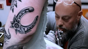 Крупный план татуировки подкова с цветком на ноге клиента и портрет мастера Бена Риффа в процессе работы