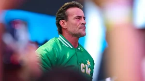 Фотография чемпиона WWE CM Пунка в зелёной спортивной куртке с белыми полосками на воротнике и золотым логотипом 'CM' со звёздой на груди. Он смотрит вправо, выражение лица сосредоточенное. Фон размыт, видны элементы стадиона или арены.