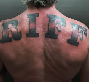 Фотография спины актера Доминика Перселла с крупной черной татуировкой RIFF, выполненной в стиле collegiate lettering