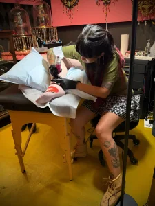 Тату-мастер Дженни Банни Дэвис в зеленой футболке и клетчатой юбке наносит татуировку на ногу клиента в своей студии в Атланте