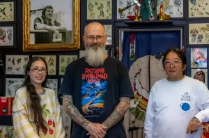 Фотография трех сотрудников легендарной тату-студии Picture Machine Tattoo в Сан-Франциско: Джен Вонг, Джейсон Стоури и Дэхи Мун, стоящих на фоне стен, украшенных старинными тату-флешами и японскими элементами декора.