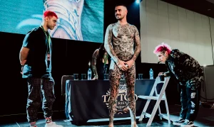 Мужчина с полностью покрытым татуировками телом стоит на сцене Australian Tattoo Expo в окружении других мастеров, демонстрируя масштабную работу в стиле blackwork и реализма.