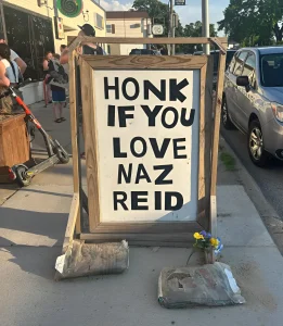 Деревянный знак на тротуаре с надписью HONK IF YOU LOVE NAZ REID, украшенный цветами и утяжеленный мешками с цементом