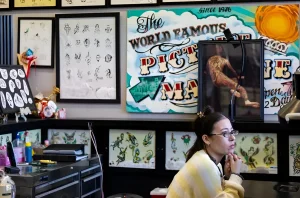 Сотрудница тату-салона Picture Machine Tattoo сидит за столом на фоне стен, украшенных винтажными плакатами и эскизами татуировок в стиле Old School
