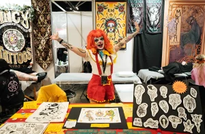 Фотография с Australian Tattoo Expo: артистка в костюме клоуна с красными волосами и татуировками на руках стоит с поднятыми руками перед стендами тату-студий и плакатами с эскизами.
