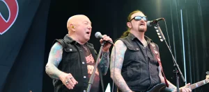 Вокалист Angry Anderson и бас-гитарист Rose Tattoo в чёрных джинсовых безрукавках с обильными цветными татуировками на руках (full sleeves) поют и играют на сцене фестиваля Skogsröjet в Швеции, 31 июля 2026