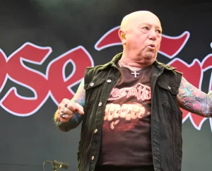 Angry Anderson, вокалист Rose Tattoo, на сцене фестиваля Skogsröjet в Швеции, 31 июля 2026; лысый мужчина в тёмной джинсовой безрукавке и футболке с логотипом группы, с цветными татуировками на обеих руках и крестом на шее, на фоне красного логотипа