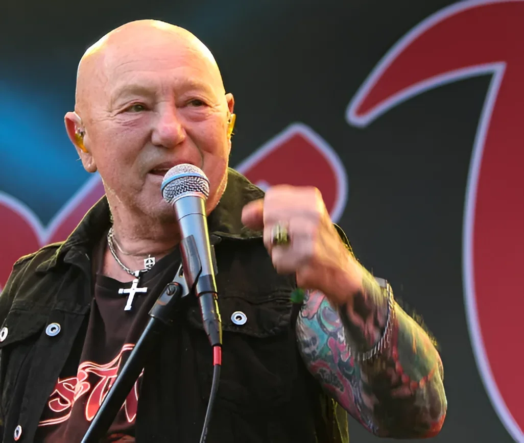 Angry Anderson, вокалист Rose Tattoo, поёт в микрофон на фестивале Skogsröjet в Швеции, указывая рукой; на поднятой руке виден яркий цветной тату-рукав в стиле old school, на шее — серебряный крест