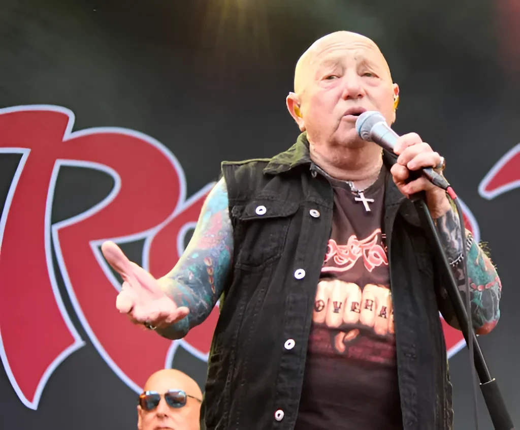 Вокалист Rose Tattoo Angry Anderson на сцене фестиваля Skogsröjet: лысый мужчина в чёрном жилете и футболке держит микрофон, на обеих руках видны крупные цветные татуировки в традиционном стиле, на шее — крест, на заднем плане красный логотип.