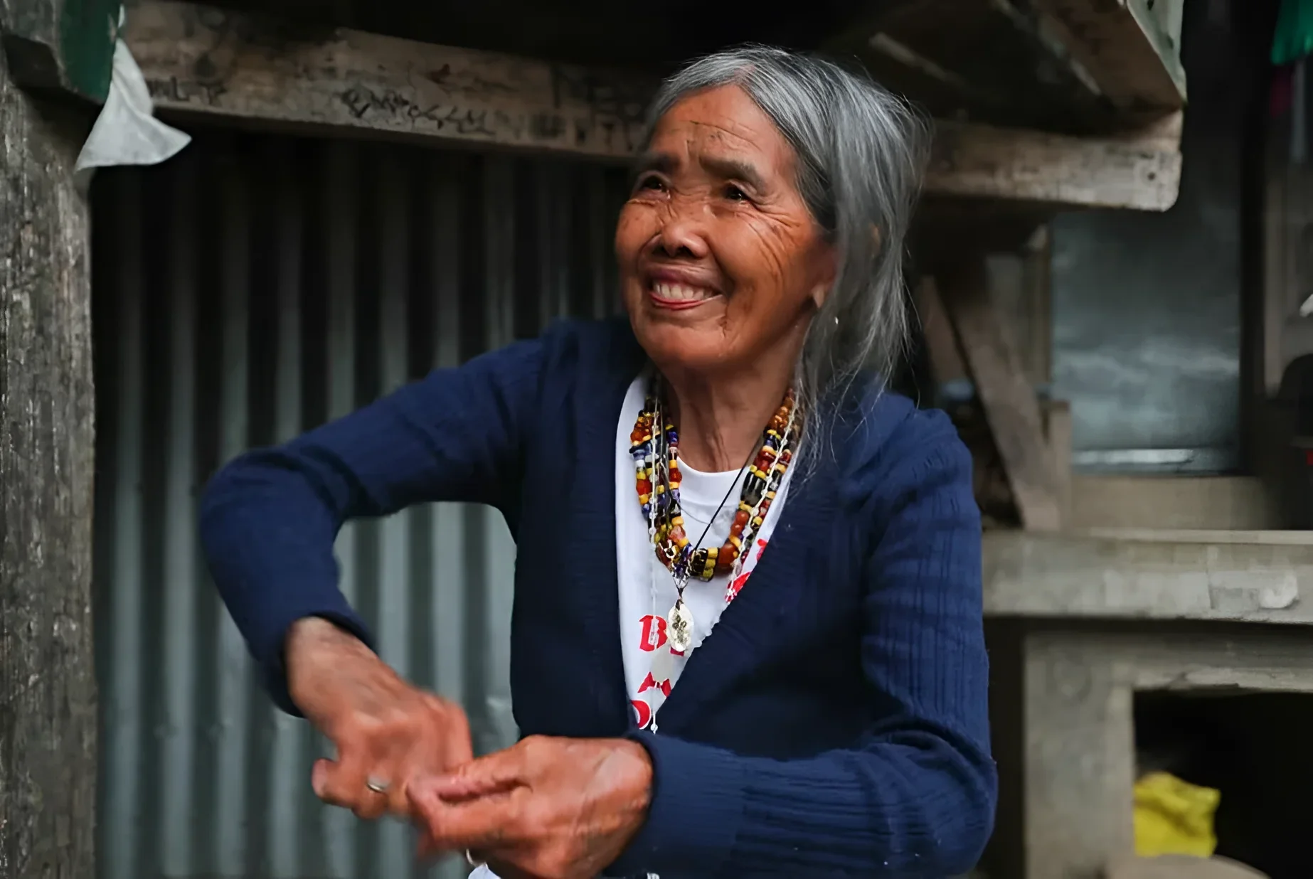 Пожилая женщина Apo Whang-Od из племени калинга улыбается, стоя у входа в дом; на ней синий кардиган, белая футболка и бусы из кораллов и бисера, на предплечье видны традиционные чёрные узоры батока
