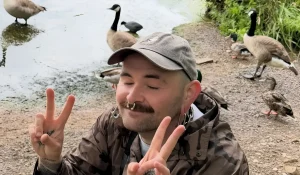 Тату-мастер Чарли Салливан в серой кепке и камуфляжной куртке делает жест мира обеими руками с татуировками на ладонях, на заднем плане водоём с канадскими гусями и другими птицами