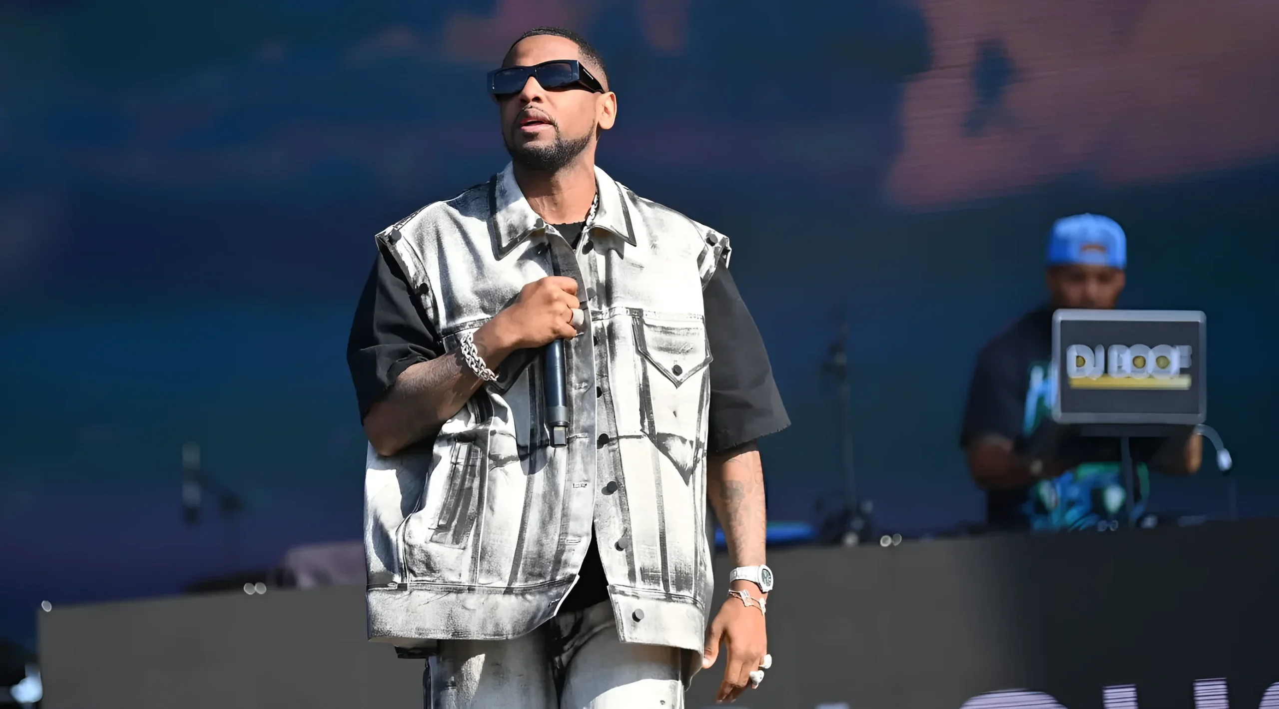 Fabolous на сцене фестиваля в солнцезащитных очках и рваной серебристой жилетке, держит микрофон; на предплечьях видны татуировки, на заднем плане диджей за пультом