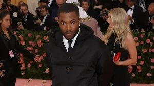 Frank Ocean в чёрном худи Prada с белым воротником и галстуком на Met Gala 2019, тема Camp, на фоне розовые цветы и фотографы