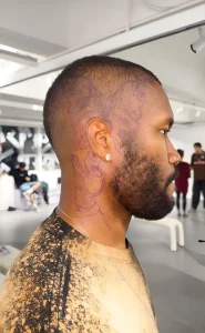Frank Ocean в профиль с фиолетовым стэнсиль-трафаретом татуировки на виске, за ухом и на шее: контур на макушке и рисунок руки с жестом, нанесённый переносной пастой перед сеансом тату.