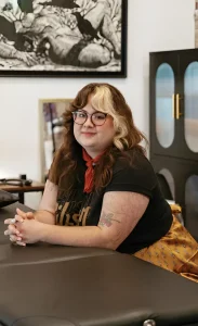 Портрет тату-художницы студии The Secret Garden Tattoo + Cosmetic Studio в Эвансвилле: женщина в очках и чёрной футболке с красным шейным платком, на предплечье видна цветная татуировка с цветком и ягодами в стиле fine line / neo-traditional