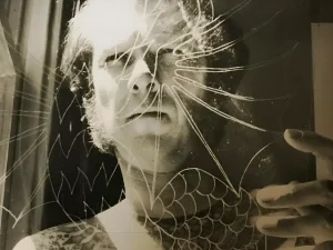 Чёрно-белый портрет гитариста Jorma Kaukonen, снятый через треснувшую стеклянную поверхность с расходящимися от центра трещинами; на плече видна плотная орнаментальная татуировка, внизу композиции — чешуйчатый узор, справа рука касается стекла.