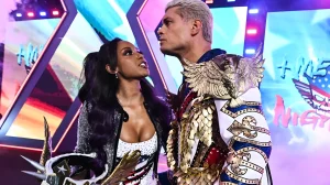 Бранди Роудс и Коди Роудс в паре на ринге WWE American Nightmare Night: Коди в белом костюме с золотым орлом на плече и чешуйчатыми элементами, на шее видна татуировка, Бранди в серебристой блестящей куртке смотрит на мужа.
