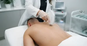 Специалист в белом халате и перчатке проводит процедуру лазерной эпиляции на спине мужчины, лежащего на кушетке в светлом кабинете, подготовка кожи перед нанесением татуировки