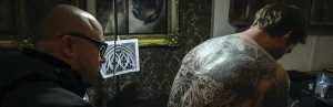 В тату-студии лысый мужчина в очках с чёрной оправой в профиль смотрит на спину другого мужчины без рубашки, на которой выполнена большая серо-чёрная татуировка в стиле neo-traditional с орнаментальными и цветочными узорами; на фоне золочёная рама с