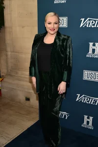 Меган Маккейн позирует на синем бэкдропе с логотипами Variety, History и USO, одетая в изумрудно-зелёный бархатный пиджак с широкими манжетами, чёрный топ и брюки того же тона, с коротким блондинистым причёсанным образом и красной помадой
