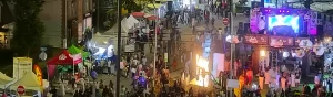 Ночной городской фестиваль Arts, Beats and Eats в Royal Oak: белые и цветные палатки вендоров, яркая сцена с экраном и световыми эффектами, декоративные фонарики и большая толпа посетителей на улице.