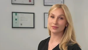 Молодая женщина со светлыми волосами в чёрной блузе и многослойной цепочке стоит в студии на фоне белой стены с четырьмя сертификатами в чёрных рамках; это украинская тату-мастер Милли Митник.