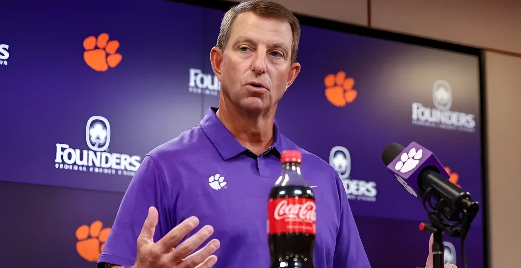 Мужчина в фиолетовой polo-рубашке с белым логотипом лапы тигра Clemson отвечает на вопросы на пресс-конференции; за ним экраны с оранжевыми логотипами Clemson и надписью Founders, справа микрофон с фирменным флагом и бутылка Coca-Cola.