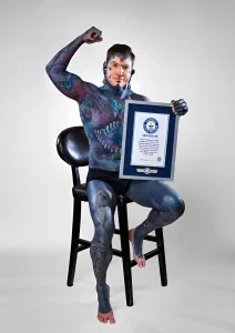 Канадец Рами Шулфилд сидит на высоком стуле, демонстрируя сертификат Guinness World Records и напряжённый бицепс; всё его тело, включая лицо, покрыто многослойными татуировками в тёмных сине-фиолетовых тонах с крупным изображением пасти на груди и