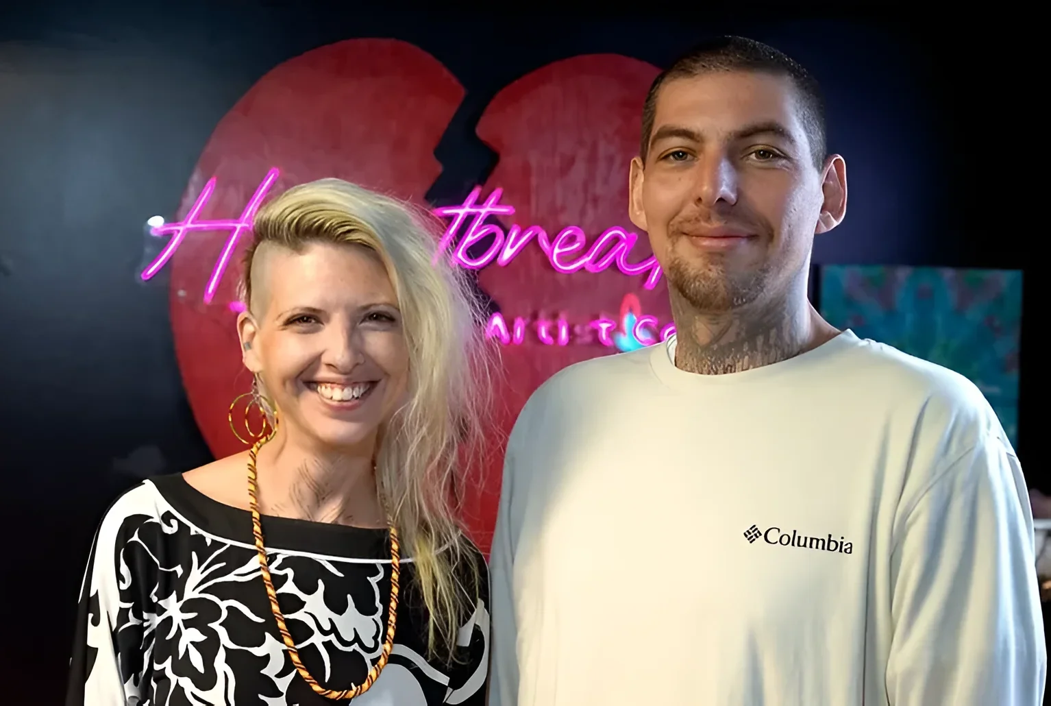 Илия Стэндиш и Тайлор Баттольф, совладельцы тату-студии Heartbreaker Tattoo and Artist Co-op, позируют на фоне неоновой вывески студии с красным сердцем; Тайлор в белой футболке Columbia с татуировкой на шее, Илия в черно-белой блузке с цветочным
