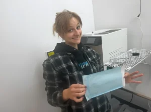 Тату-мастер Stella Johnson улыбается и держит в руках стерильный пакет для автоклавирования на фоне белого автоклава и металлической сетчатой полки для инструментов в студии боди-арта в Alpena.
