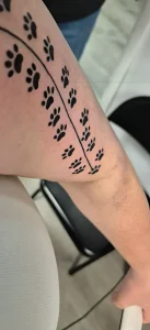 Чёрно-белая татуировка в стиле blackwork на внутренней стороне предплечья: две параллельные линии, между которыми по диагонали выстроены лапки кошек и собак, символизирующие 40 питомцев, которых любила хозяйка за 50 лет