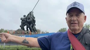 Ветеран морского пехоты США Том Кеннеди в серой кепке с эмблемой Marine Corps вытягивает предплечье с чёрно-белой татуировкой мемориала «Подъём флага на Иводзиме» на фоне оригинальной скульптуры мемориала Корпуса морской пехоты в Вашингтоне