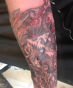 Детальный снимок татуировки в японском стиле irezumi на теле Ронана Кийтинга: дракон с чешуей, раскрытый веер, ветви сакуры, горы и солнце в чёрно-серой гамме с красными акцентами