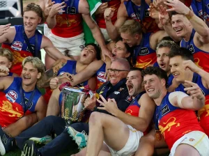 Команда Brisbane Lions в сине-красных майках с логотипом льва празднует победу, поднимая кубок AFL Premiership; на руках и ногах игроков видны татуировки
