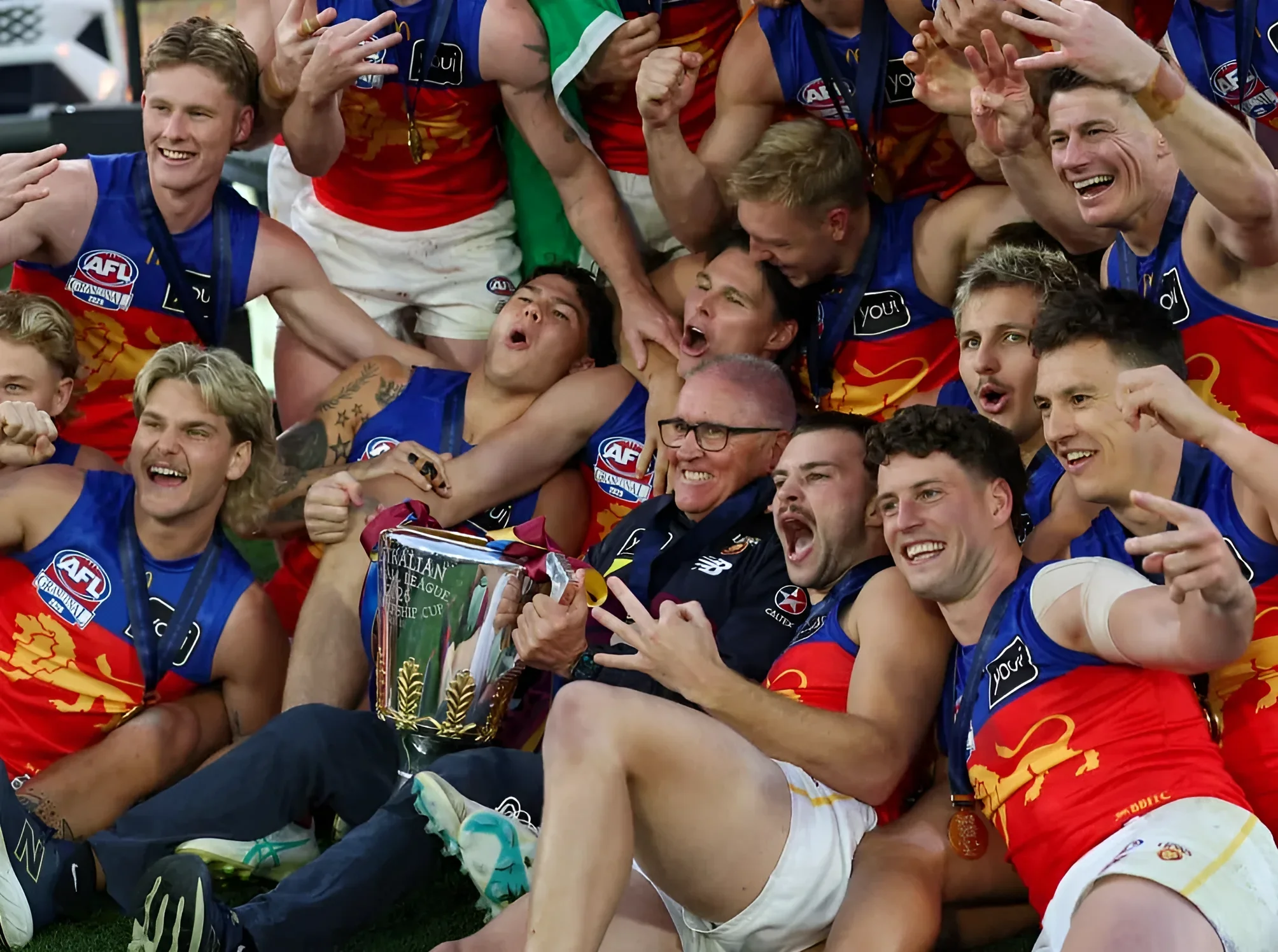 Команда Brisbane Lions в сине-красных майках с логотипом льва празднует победу, поднимая кубок AFL Premiership; на руках и ногах игроков видны татуировки