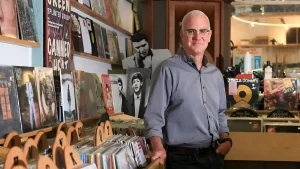 Джефф Лейпер в серой рубашке и очках стоит в виниловом магазине The Record Centre на Веллингтон-стрит, на фоне стеллажей с пластинками и постеров Green Day, Canned Heat, Элвиса Пресли и The Beatles
