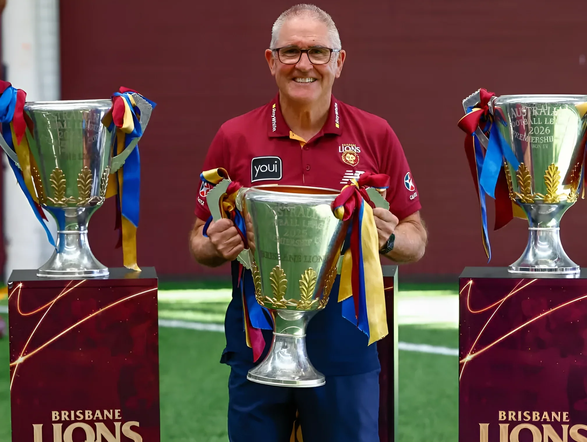 Крис Фэгэн в бордовой polo-рубашке Brisbane Lions держит серебряный трофей с золотыми листьями и цветной лентой, рядом на подиумах стоят ещё два кубка клуба