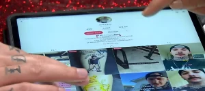 Рука с чёрными татуировками на пальцах держит планшет, на экране которого открыт профиль TikTok artbydudus из Оклахомы с 170,6K подписчиков; палец указывает на сетку видео с татуировками, на фоне красный глиттер