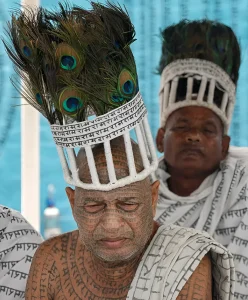 Двое мужчин из общины Рамнами с чёрными татуировками повторяющихся надписей на деванагари на лицах, лысых макушках, шее и плечах; на головах белые венцы с павлиньими перьями и лентами с текстом, на плечах белые ткани с напечатанным текстом, на фоне