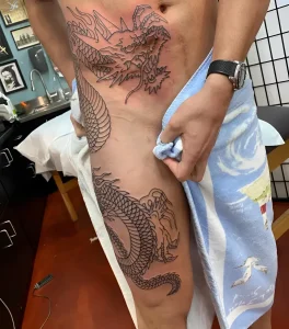 Свежая контурная татуировка японского дракона в технике blackwork, идущая от грудной клетки по правому боку и бедру; мастерская, полотенце, этап line work