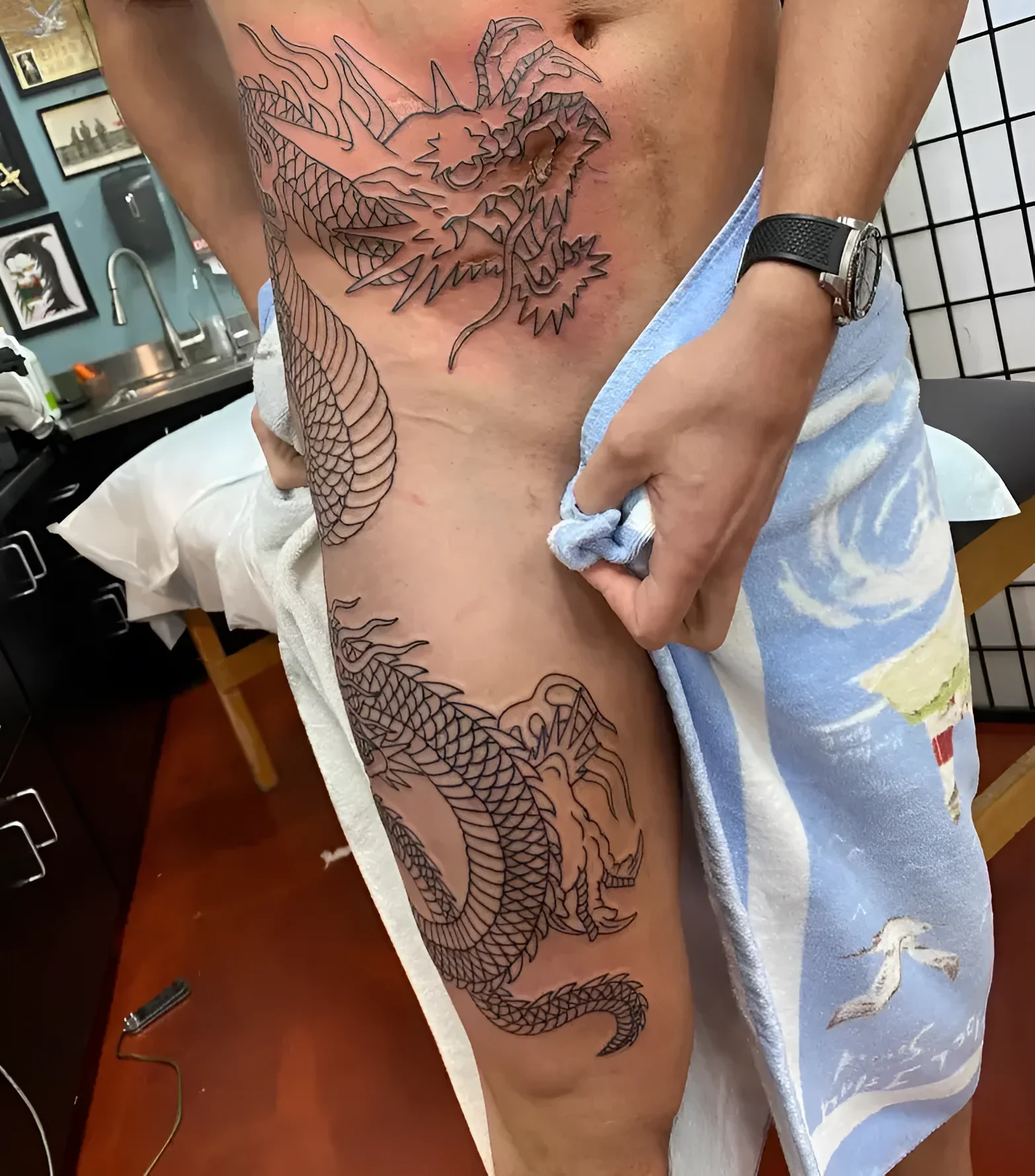 Свежая контурная татуировка японского дракона в технике blackwork, идущая от грудной клетки по правому боку и бедру; мастерская, полотенце, этап line work