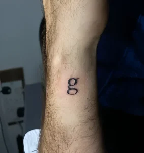 Минималистичная чёрная контурная татуировка строчной буквы «g» в типографическом стиле на волосатом предплечье Дэвида Теннанта