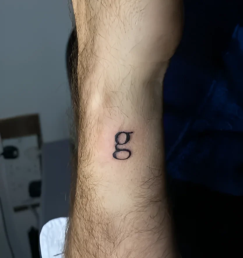 Минималистичная чёрная контурная татуировка строчной буквы «g» в типографическом стиле на волосатом предплечье Дэвида Теннанта