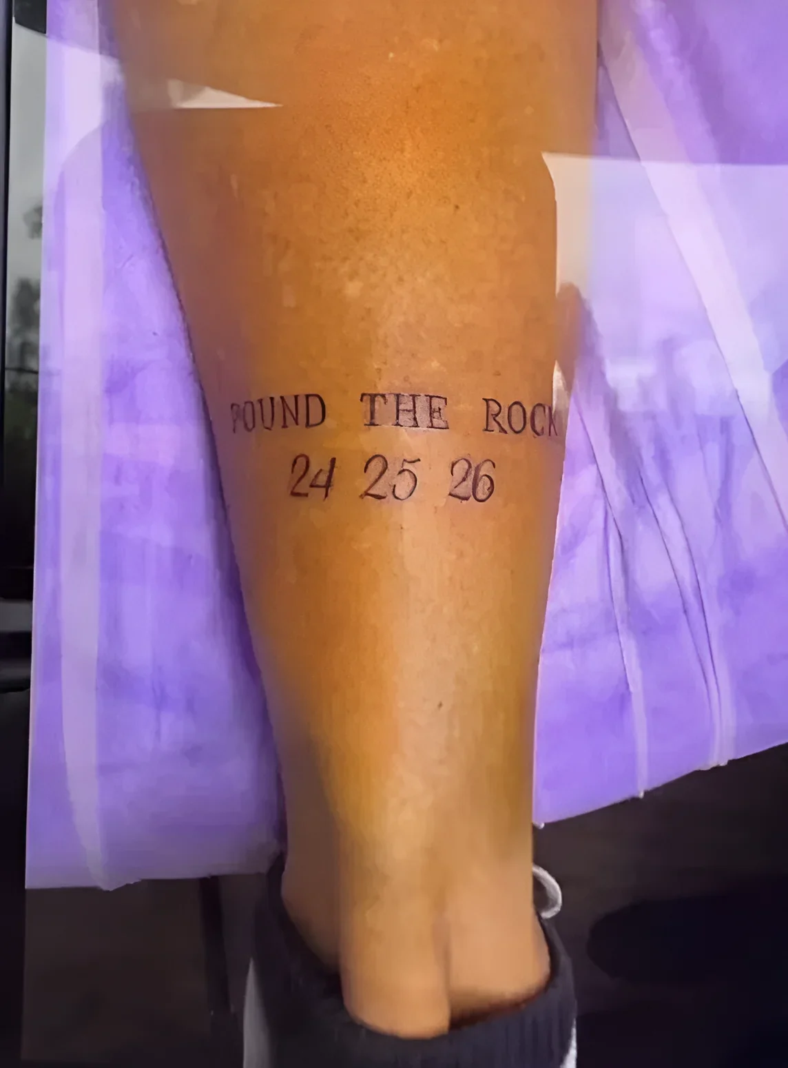 Чёрная татуировка в стиле lettering с надписью POUND THE ROCK и цифрами 24 25 26 на передней поверхности голени Криса Фэгэна на фоне фиолетовой ткани Brisbane Lions