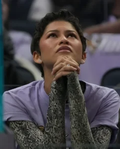 Зендея на трибунах арены Chase Center в Сан-Франциско во время финала WNBA: короткая тёмная стрижка, руки сложены в замок у подбородка, на ней лаваандовая футболка поверх длинных рукавов с плотным чёрно-белым змеиным принтом, имитирующим чешую.