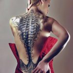 Фото татуировки на спине женщины, изображающей корсет, состоящий из костей и металлических деталей. Реалистичный стиль, эффект обнаженной кожи.